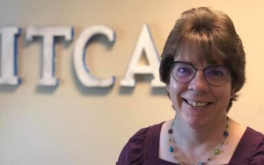 A white woman with short hair and glasses poses in front of an ITCA sign.