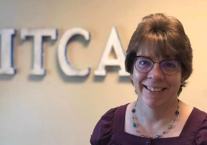 A white woman with short hair and glasses poses in front of an ITCA sign.